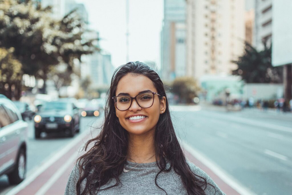 pexels-photo-1239291-1239291 A young woman with glasses smiling on a city street, embracing urban lifestyle.